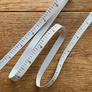 May include: Two white measuring tapes with black markings on a wooden surface. The tapes are in a crisscross pattern, with one tape forming a loop.