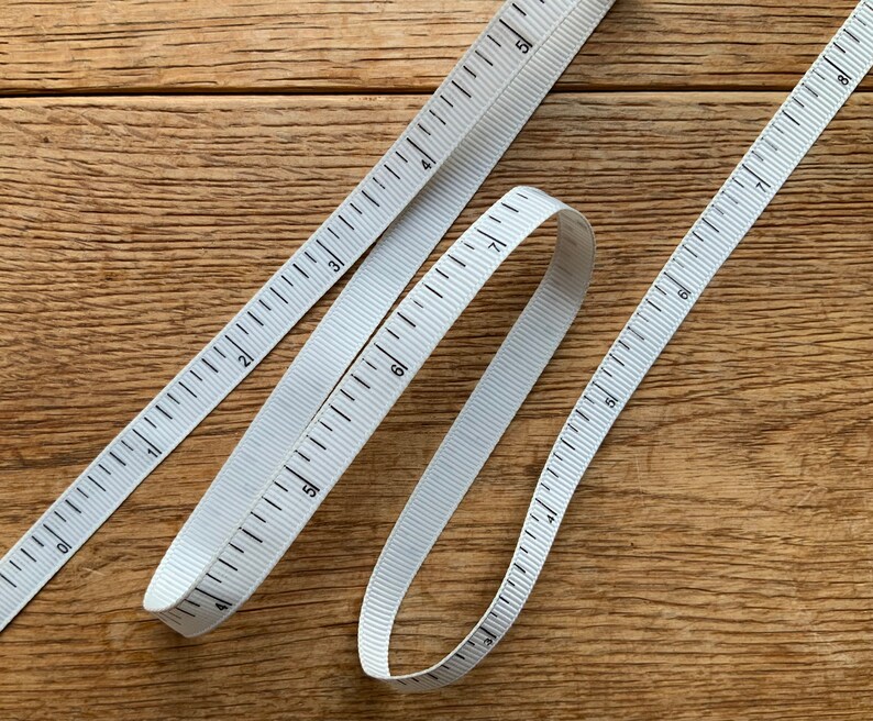 May include: Two white measuring tapes with black markings on a wooden surface. The tapes are in a crisscross pattern, with one tape forming a loop.