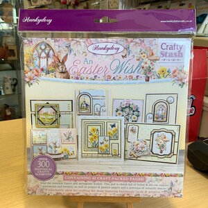 May include: A craft paper pad with an Easter theme. The cover features a colourful illustration of a church, flowers, and a bunny rabbit. The title "An Easter Wish" is printed in a decorative font. The pad contains 42 craft-packed pages and over 300 individual crafty elements.