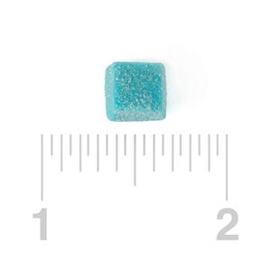May include: A single, square, light blue gummy candy with a textured, sugar-coated surface. The candy is positioned on a white background next to a ruler, with measurements from 1 to 2 inches.