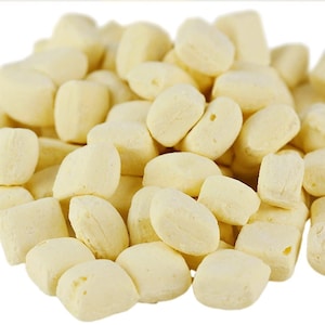 May include: A pile of small, pale yellow, irregularly shaped candies. The candies have a slightly rounded or rectangular form, with a matte finish. Some pieces have small holes. The overall appearance suggests a type of confectionery or snack food.