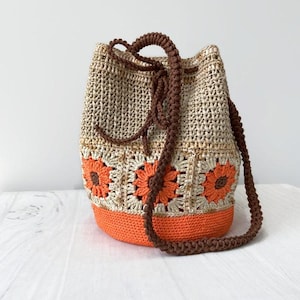 May include: A handmade crochet bucket bag in beige and orange. The bag features a brown drawstring closure and strap. The lower section and floral details are orange, with a beige body. The bag is designed with a sunflower pattern.