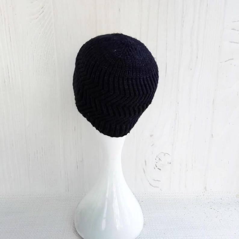 May include: A black knit beanie with a chevron pattern. The hat is displayed on a white mannequin head against a white wooden background. The beanie is made of a soft, textured material.