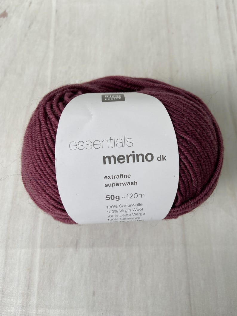 May include: A ball of burgundy-colored merino wool yarn with a white label. The label reads "essentials merino dk extrafine superwash" and lists the weight as 50g ~120m. The yarn is 100% virgin wool.
