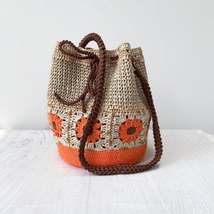 May include: A handmade crochet bucket bag with a drawstring closure. The bag features a beige body with orange floral accents and a solid orange base. It has a brown braided strap and drawstring. The bag is designed for carrying items.