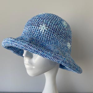 Puede incluir: Un sombrero azul tejido a ganchillo con ala ancha y diseño floral. El sombrero es una mezcla de hilos azules claros y oscuros, con pequeñas flores bordadas en azul claro. El sombrero se exhibe en una cabeza de maniquí blanca.