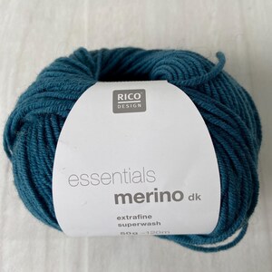 May include: A ball of teal-colored yarn with a white label. The label reads "RICO DESIGN essentials merino dk extrafine superwash 50g - 120m". The yarn is tightly wound and appears soft.