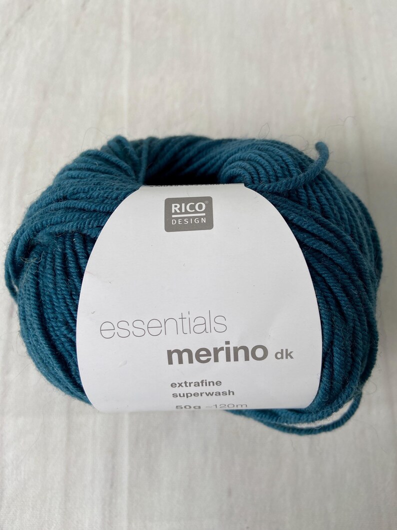 May include: A ball of teal-colored yarn with a white label. The label reads "RICO DESIGN essentials merino dk extrafine superwash 50g - 120m". The yarn is tightly wound and appears soft.