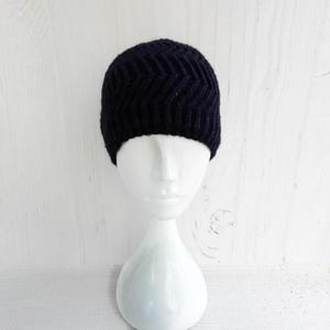 May include: A dark navy blue knit beanie with a chevron pattern. The hat is displayed on a white mannequin head against a white wooden background. The beanie is a winter accessory.