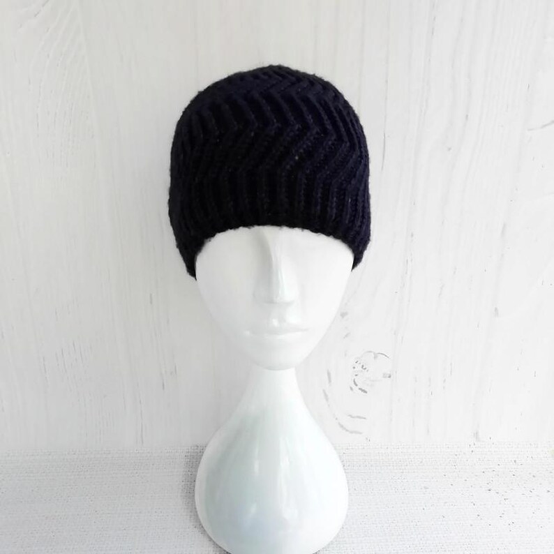 May include: A dark navy blue knit beanie with a chevron pattern. The hat is displayed on a white mannequin head against a white wooden background. The beanie is a winter accessory.