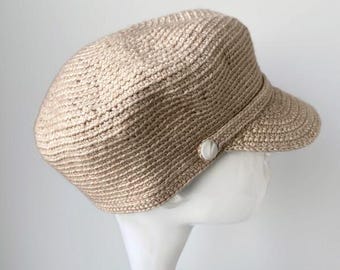 May include: A beige newsboy cap crafted from a textured, woven material. The hat features a rounded crown, a short stiff brim, and a small, circular white button detail positioned on the side where the brim meets the main body of the cap.