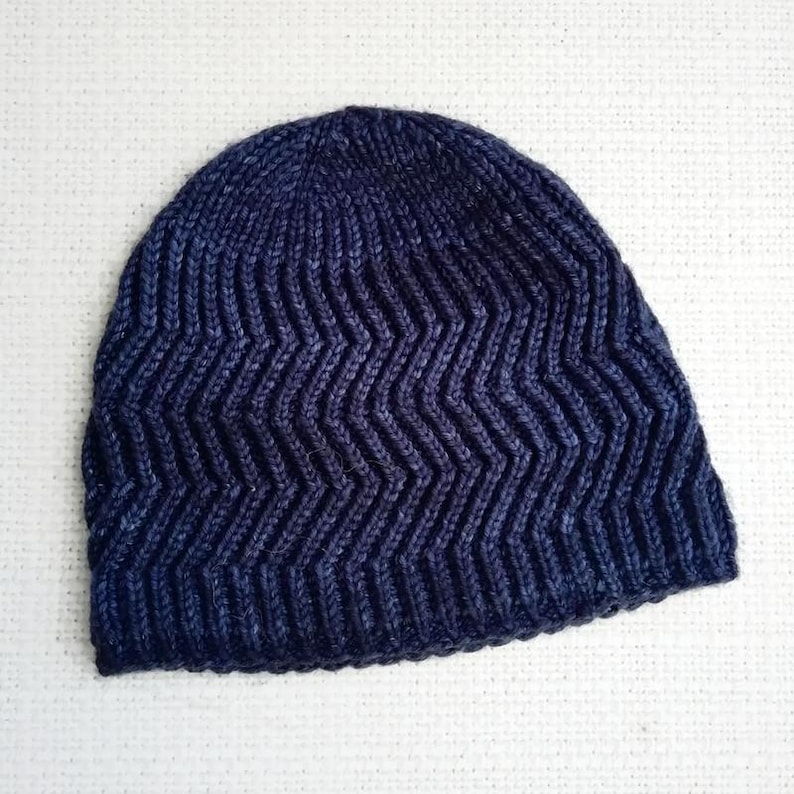 May include: A navy blue knit beanie with a chevron pattern. The hat is made of a textured yarn and has a ribbed brim. The beanie is a winter accessory.