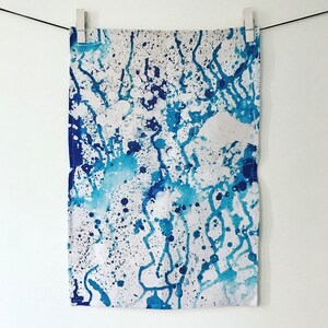 Puede incluir: Un paño de cocina blanco con un diseño abstracto de pintura salpicada azul y blanca. El diseño presenta salpicaduras y rayas de pintura azul y blanca.