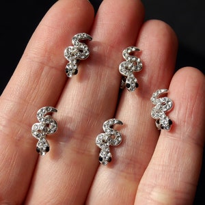 May include: Five silver snake-shaped earrings with clear rhinestones. The earrings are on a hand with a black background.
