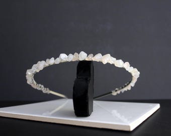 May include: A delicate, handcrafted headband featuring a series of small, irregularly shaped, light-colored stones. The stones are strung along a thin, silver-toned wire, creating a crown-like appearance. The headband is displayed on a black stand against a dark background.