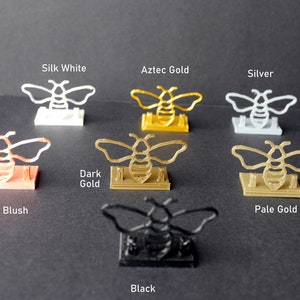 May include: A set of seven bee-shaped phone stands in various colors, including silk white, Aztec gold, silver, rose blush, dark gold, pale gold, and black.