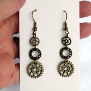 May include: A pair of steampunk earrings. Each earring has three bronze-coloured gear-shaped charms hanging from a hook. The gears are different sizes, with the largest at the bottom. They are displayed on a white card.