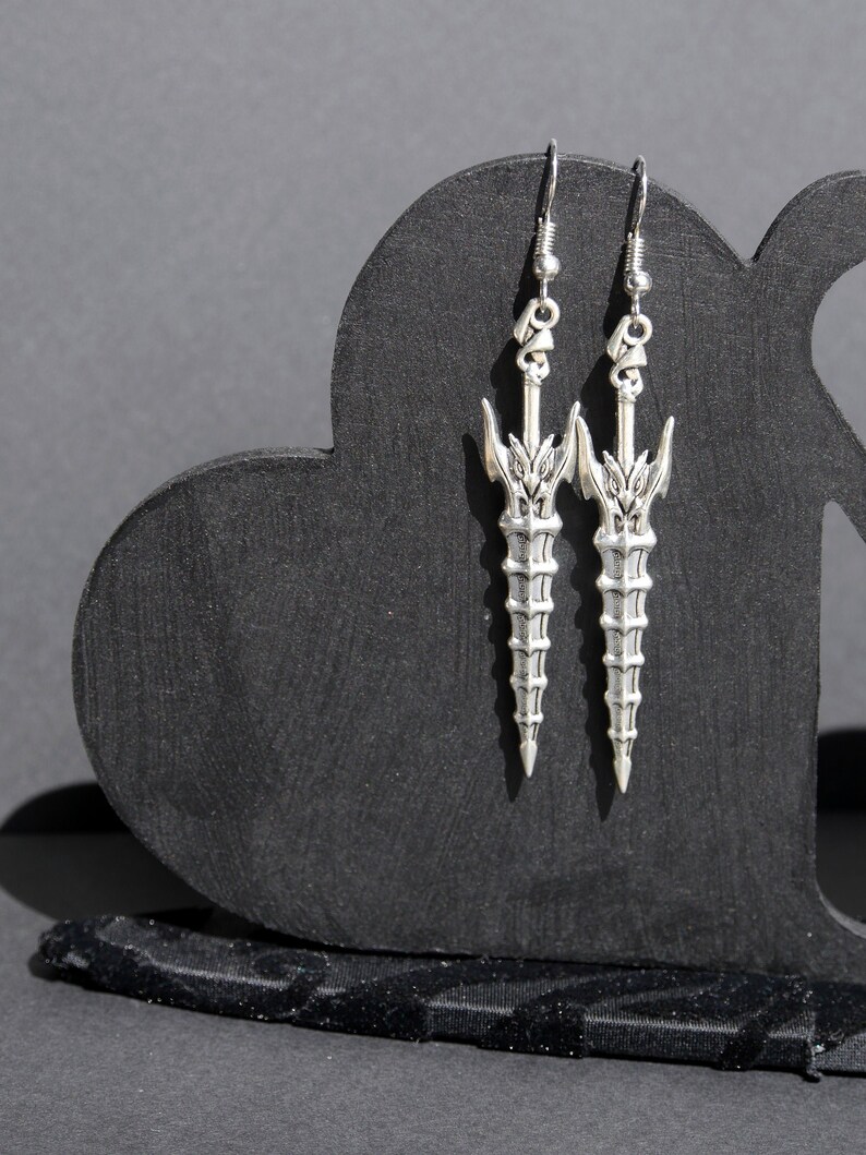 May include: A pair of silver earrings shaped like swords with intricate details. The earrings are hanging from a black heart-shaped display stand.