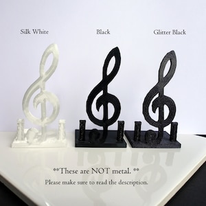 May include: Three 3D printed treble clef music note holders in different colours: silk white, black, and glitter black. The text "These are NOT metal." is printed below the holders.