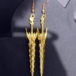 May include: A pair of gold-tone earrings shaped like swords with intricate details. The earrings are hanging from a black display stand.