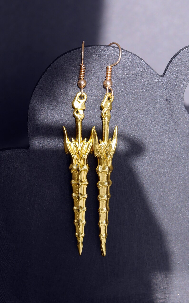 May include: A pair of gold-tone earrings shaped like swords with intricate details. The earrings are hanging from a black display stand.