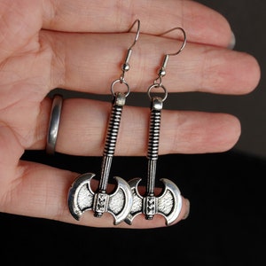 May include: A pair of silver axe earrings with a detailed design. The earrings are hanging from silver hooks.