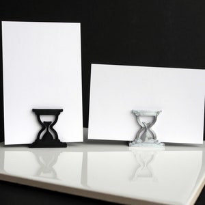 May include: Two hourglass-shaped card holders, one black and one silver, holding blank white cards.