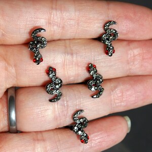 May include: Four pairs of black snake earrings with red rhinestone eyes. The earrings are on a hand.