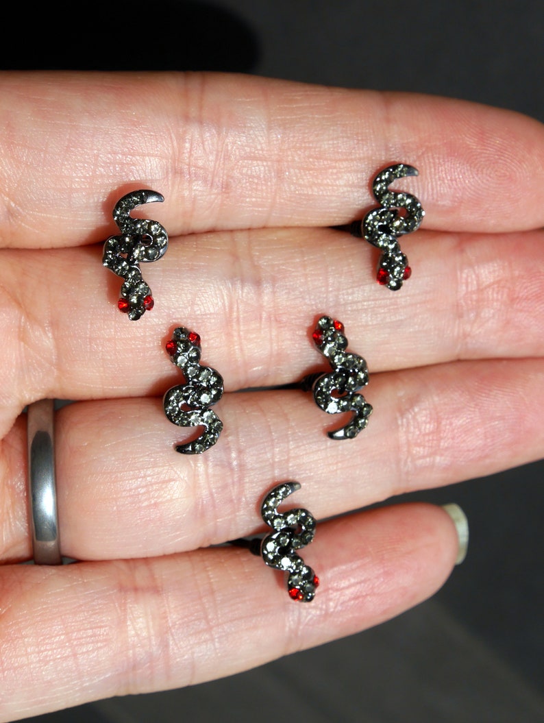 May include: Four pairs of black snake earrings with red rhinestone eyes. The earrings are on a hand.