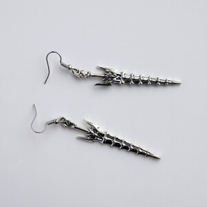 May include: A pair of silver earrings shaped like swords with intricate details. The swords have a skeletal design and are attached to silver hooks.