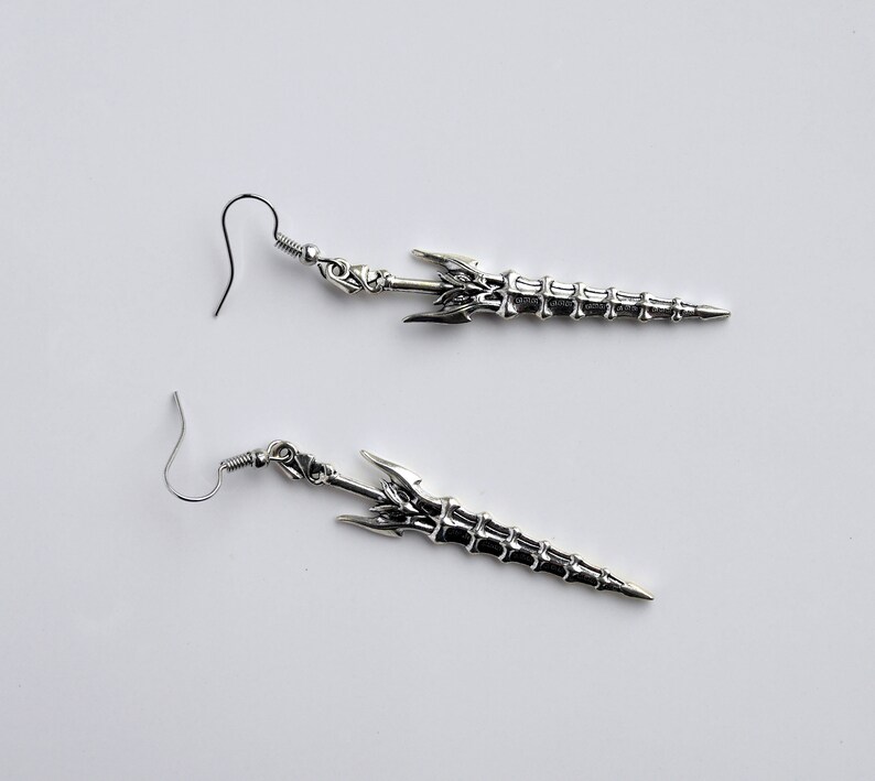 May include: A pair of silver earrings shaped like swords with intricate details. The swords have a skeletal design and are attached to silver hooks.