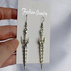 May include: A pair of silver sword-shaped dangle earrings with a detailed design. The earrings are on a white card with the text "Fashion Jewelry".