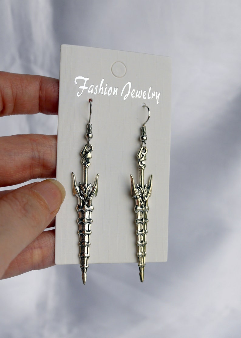 May include: A pair of silver sword-shaped dangle earrings with a detailed design. The earrings are on a white card with the text "Fashion Jewelry".