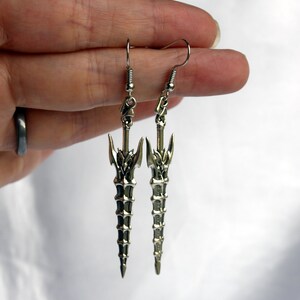 May include: A pair of silver earrings featuring a detailed sword design. The swords are long and slender with a pointed tip and intricate details.