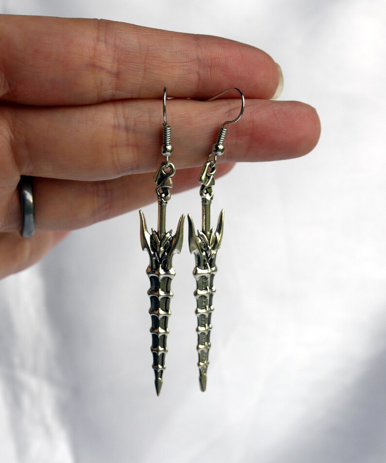 May include: A pair of silver earrings featuring a detailed sword design. The swords are long and slender with a pointed tip and intricate details.
