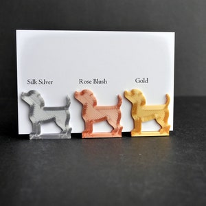 May include: Three dog-shaped figures in silk silver, rose blush, and gold colors are displayed against a white card. Each figure stands on a rectangular base. The text Silk Silver, Rose Blush, and Gold is printed above each respective dog figure.