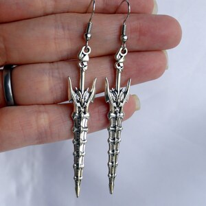May include: A pair of silver earrings shaped like swords with intricate details. The swords have a pointed tip and a decorative design on the hilt.