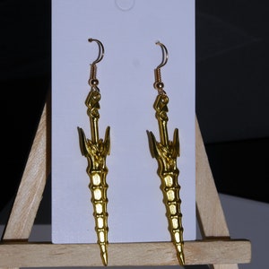 May include: A pair of gold-colored earrings shaped like swords with intricate details. The earrings are hanging from a white card.