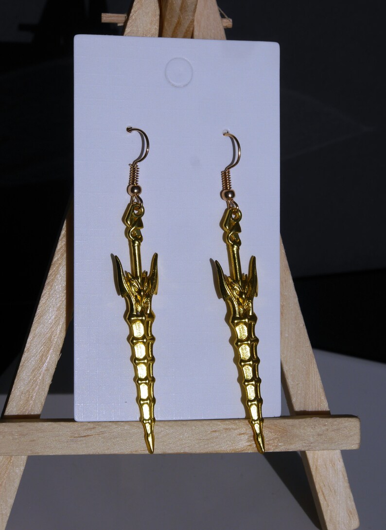 May include: A pair of gold-colored earrings shaped like swords with intricate details. The earrings are hanging from a white card.