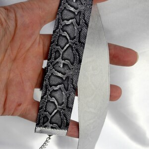 May include: A black and white snake skin patterned choker necklace with a silver chain and clasp. The choker is made of fabric and is shown folded in half.