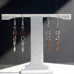 May include: Four steampunk earrings on a white stand. Each earring has a silver hook, chain, and gear details. Two earrings have copper-coloured arrow pendants, and two have silver pendants. The gears are silver and copper-toned.