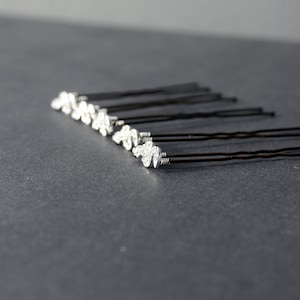 May include: A set of ten silver hair pins with decorative swirls on the ends. The pins are arranged in a row on a gray surface.