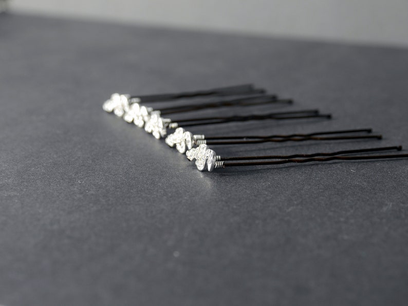 May include: A set of ten silver hair pins with decorative swirls on the ends. The pins are arranged in a row on a gray surface.