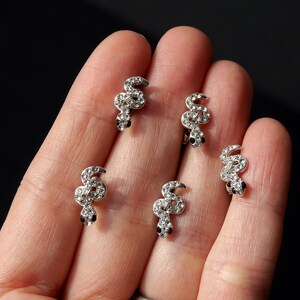 May include: Five silver snake-shaped earrings with clear rhinestones. The earrings are on a hand.