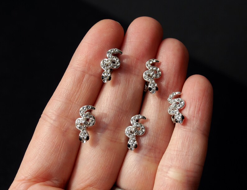 May include: Five silver snake-shaped earrings with clear rhinestones. The earrings are on a hand.