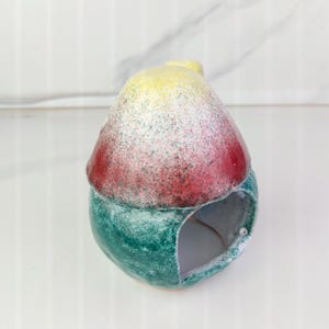 K&ouml;nnte beinhalten: Ein Keramik-Vogelhaus mit abgerundeter Oberseite und einer kreisf&ouml;rmigen &Ouml;ffnung. Die Oberseite ist mit gelber, roter und wei&szlig; gesprenkelter Farbe bemalt. Der Boden ist gr&uuml;n mit gesprenkelter Oberfl&auml;che. Das Vogelhaus ist als Dekorationsartikel konzipiert.
