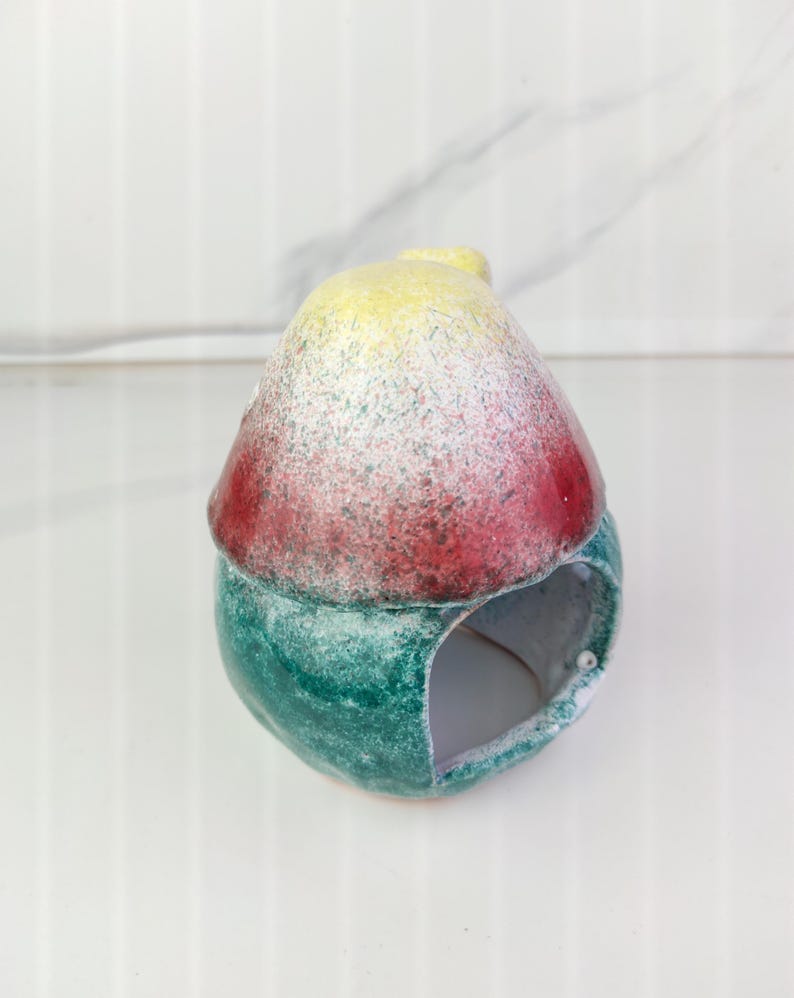 K&ouml;nnte beinhalten: Ein Keramik-Vogelhaus mit abgerundeter Oberseite und einer kreisf&ouml;rmigen &Ouml;ffnung. Die Oberseite ist mit gelber, roter und wei&szlig; gesprenkelter Farbe bemalt. Der Boden ist gr&uuml;n mit gesprenkelter Oberfl&auml;che. Das Vogelhaus ist als Dekorationsartikel konzipiert.