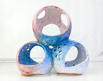 May include: A ceramic sculpture composed of three interconnected spherical forms with large circular openings. The surface features a speckled glaze transitioning from shades of pink and red at the top to vibrant blue and white at the base.