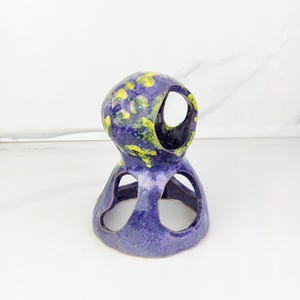 May include: A handcrafted ceramic sculpture in shades of blue and purple, adorned with yellow accents. The unique piece features open circular cutouts and a textured surface, showcasing an artistic, abstract design.