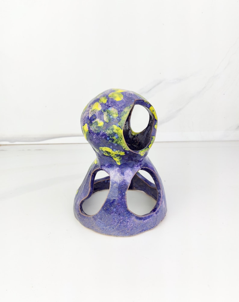 May include: A handcrafted ceramic sculpture in shades of blue and purple, adorned with yellow accents. The unique piece features open circular cutouts and a textured surface, showcasing an artistic, abstract design.